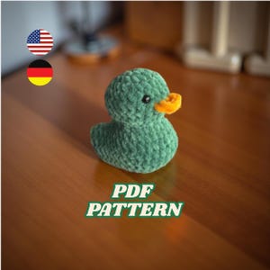 May include: A handmade green crochet duck with an orange beak and black eyes. The duck is sitting on a wooden surface. The text "PDF PATTERN" is displayed in green and white.