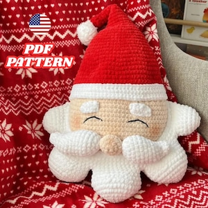 Puede incluir: Un cojín de ganchillo de Papá Noel con gorro rojo y barba blanca. El cojín tiene una cara sonriente y está sobre una manta roja estampada. El texto "PDF PATTERN" es visible en la imagen.