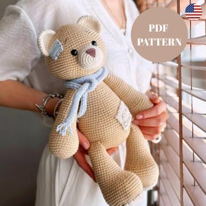 May include: A handmade, beige crochet teddy bear with a light blue scarf and a small blue patch. The bear has a stitched nose and eyes. The image includes a circular graphic with the text "PDF PATTERN".