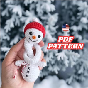 May include: A crocheted snowman with a red hat, black eyes, and an orange nose. The snowman has a white body and a scarf. The text "PDF PATTERN" is visible in the image. The snowman is a handmade item.