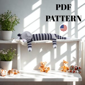 May include: A crocheted cat in gray and navy blue stripes is lying on a white shelf. The cat has a gray head, pink nose, and white paws. The text "PDF PATTERN" is above the cat, with an American flag graphic.