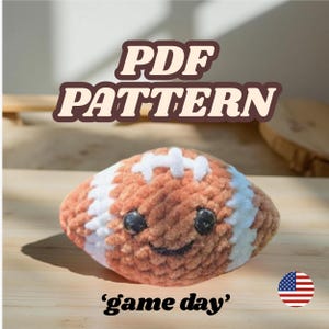 以下が含まれることがあります： 笑顔と白いストライプの、かぎ針編みのフットボール。テキスト「PDF PATTERN」がフットボールの上に表示され、「game day」が下に書かれています。アメリカの国旗が右下にあります。