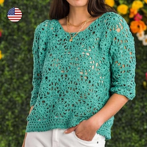 May include: A teal crochet top with a floral pattern. The top has a scoop neck and three-quarter sleeves. The crochet design creates an open, airy texture. The person is wearing a gold necklace and white pants. The background is a green hedge with yellow flowers.