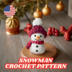 Puede incluir: Un muñeco de nieve tejido a ganchillo con gorro y bufanda burdeos, ojos de botón negros y nariz de zanahoria naranja. El muñeco de nieve está sobre una base de madera. El texto "SNOWMAN CROCHET PATTERN" está en la parte inferior.