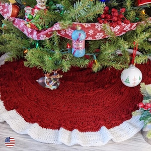 Pode incluir: Uma saia de árvore de Natal feita à mão em vermelho escuro com uma borda branca ondulada. A saia é de crochê e tem um padrão texturizado. Ela envolve uma árvore de Natal decorada com enfeites e luzes.