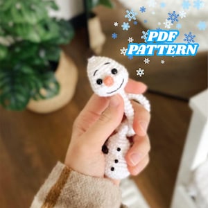 Könnte beinhalten: Ein kleiner, weiß gehäkelter Schneemann mit Karottennase, schwarzen Augen und einem lächelnden Mund. Der Schneemann hat einen runden Kopf und einen Körper mit schwarzen Knöpfen. Der Text "PDF PATTERN" ist im Bild enthalten.