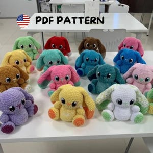 May include: A group of colourful, handmade crochet bunny plushies. The bunnies are in various colours, including green, red, blue, pink, yellow, brown, and white. The text "PDF PATTERN" is displayed above the bunnies. These plushies are sitting on a white surface.