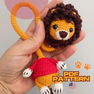 May include: Crocheted lion toy with a yellow body, red shirt, and brown mane. The lion has a friendly face with black eyes and a brown nose. The toy includes a loop and the text "PDF PATTERN".