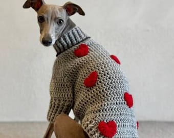 Op de afbeelding: Een grijze gehaakte hondentrui met rode hartapplicaties. De trui heeft een coltrui en wordt gedragen door een grijze en bruine Italiaanse Greyhound. De hond zit op een tapijtoppervlak.
