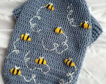 May include: A blue crocheted dog sweater with embroidered bees and white stitching.
