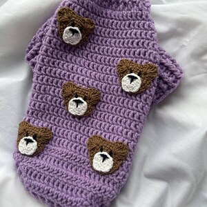 Op de afbeelding: Een lavendelgehaakte hondentrui met bruine berenapplicaties. De trui heeft een coltrui-ontwerp en is versierd met vijf berengezichten, elk met witte en zwarte details. Handgemaakte dierenkleding.