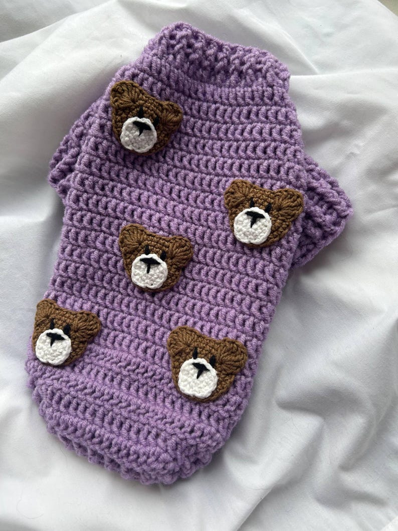 Op de afbeelding: Een lavendelgehaakte hondentrui met bruine berenapplicaties. De trui heeft een coltrui-ontwerp en is versierd met vijf berengezichten, elk met witte en zwarte details. Handgemaakte dierenkleding.