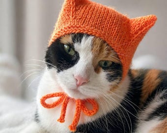 Op de afbeelding: Een lapjeskat draagt een oranje gebreide hoed met puntige oren en een bijpassende strik. De kat heeft groene ogen en een roze neus. De hoed en strik zijn fel oranje van kleur, wat contrasteert met de zwart-witte en oranje vacht van de kat.