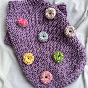Op de afbeelding: Een paarse gehaakte hondentrui versierd met kleurrijke donut-appliqu&eacute;s. De trui heeft een geribbelde kraag en is versierd met donuts in roze, geel, groen en blauw met witte hagelslag.