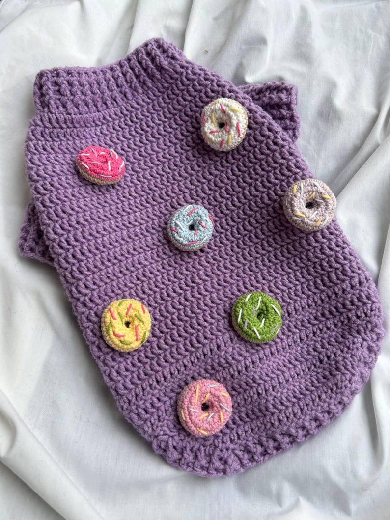 Op de afbeelding: Een paarse gehaakte hondentrui versierd met kleurrijke donut-appliqu&eacute;s. De trui heeft een geribbelde kraag en is versierd met donuts in roze, geel, groen en blauw met witte hagelslag.
