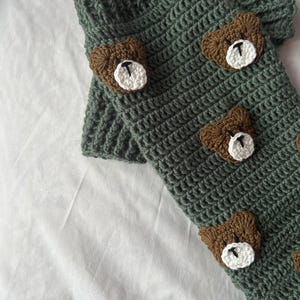 Op de afbeelding: Handgehaakte beenwarmers in een gedempte groene kleur, versierd met bruine berenapplicaties met witte en zwarte details. De beenwarmers zijn gemaakt met een getextureerde steek, perfect om een vleugje speelsheid aan elke outfit toe te voegen.