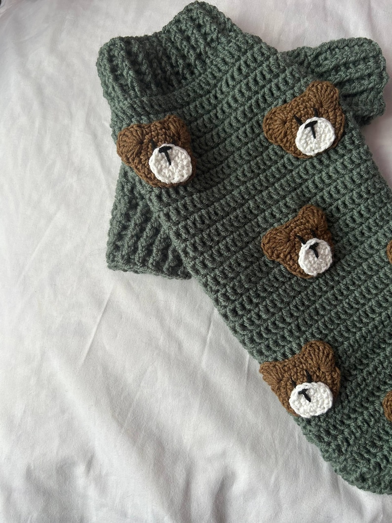 Op de afbeelding: Handgehaakte beenwarmers in een gedempte groene kleur, versierd met bruine berenapplicaties met witte en zwarte details. De beenwarmers zijn gemaakt met een getextureerde steek, perfect om een vleugje speelsheid aan elke outfit toe te voegen.