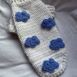 Op de afbeelding: Een witte gehaakte sok met vijf blauwe wolkenapplicaties. De sok is handgemaakt met een getextureerde steek en heeft een geribbelde boord. De wolken zijn een donkerdere tint blauw en geven het ontwerp een speelse uitstraling.
