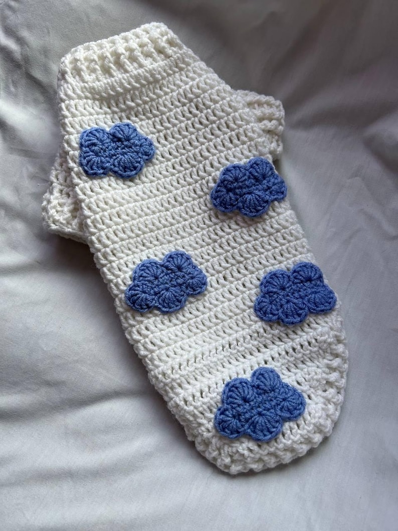 Op de afbeelding: Een witte gehaakte sok met vijf blauwe wolkenapplicaties. De sok is handgemaakt met een getextureerde steek en heeft een geribbelde boord. De wolken zijn een donkerdere tint blauw en geven het ontwerp een speelse uitstraling.