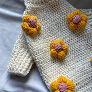 Op de afbeelding: Cr&egrave;mekleurige gehaakte trui versierd met felgele en roze bloemenapplicaties. De trui heeft een gestructureerd steekpatroon en lange mouwen. De bloemen zijn gelijkmatig verdeeld over de voorkant van het kledingstuk, wat een speelse touch geeft.