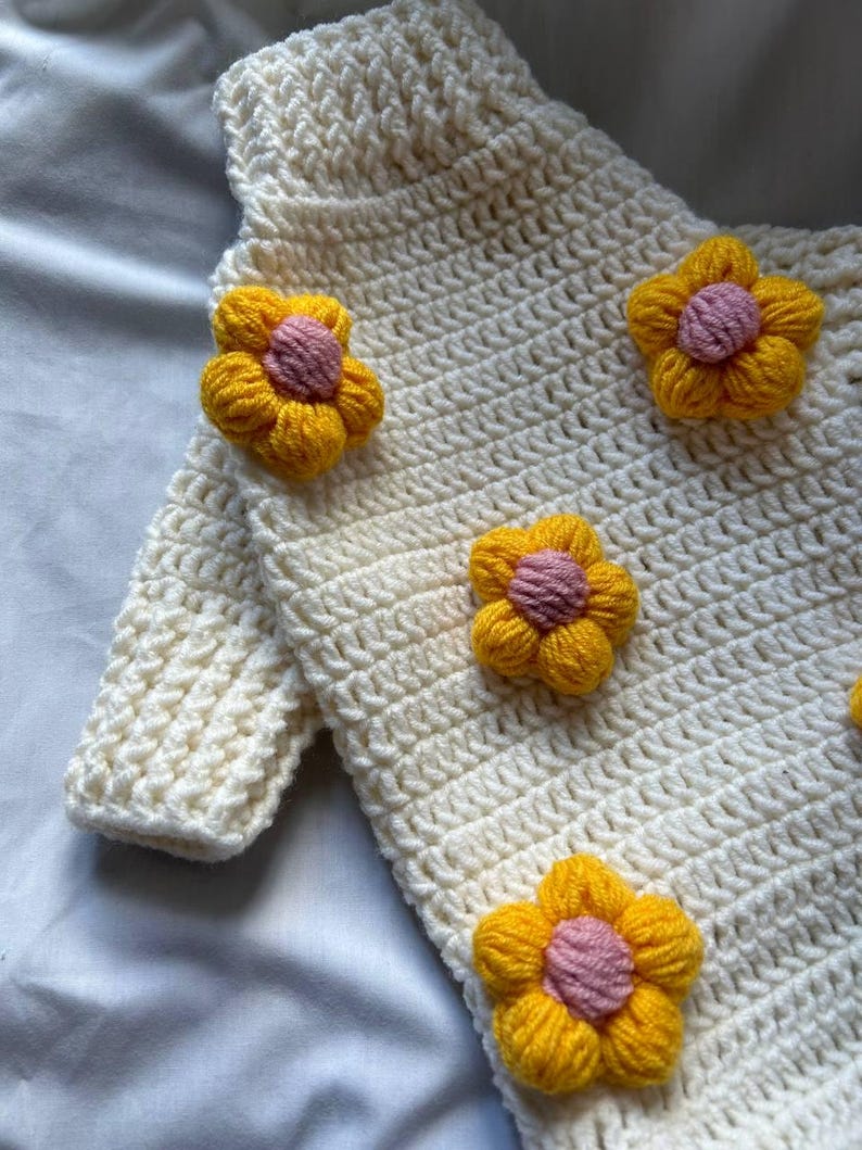 Op de afbeelding: Cr&egrave;mekleurige gehaakte trui versierd met felgele en roze bloemenapplicaties. De trui heeft een gestructureerd steekpatroon en lange mouwen. De bloemen zijn gelijkmatig verdeeld over de voorkant van het kledingstuk, wat een speelse touch geeft.
