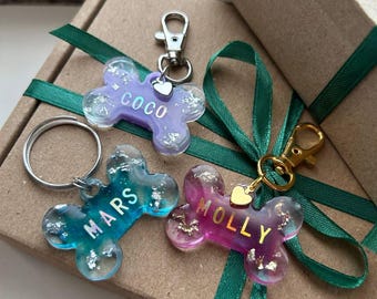 May include: Three personalized dog bone-shaped keychains in shades of blue, purple, and pink. Each keychain has a name in white lettering and silver or gold flakes. They are attached to silver and gold clasps, and are presented on a brown box with green ribbon.