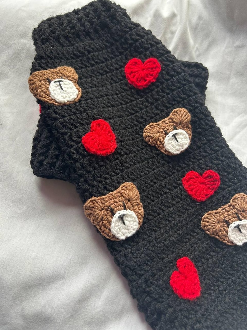 Op de afbeelding: Zwarte gehaakte mouw met applicaties van bruine teddyberen en rode harten. De teddyberen hebben witte gezichten en zijn op de mouw genaaid. De harten zijn rood en zijn ook op de mouw genaaid. De mouw is gemaakt van een getextureerd, gebreid materiaal.