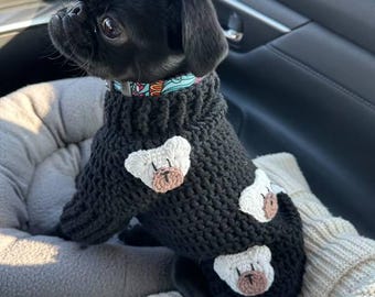 May include: A black pug wearing a black knitted sweater with white bear appliques. The dog is sitting in a car seat.