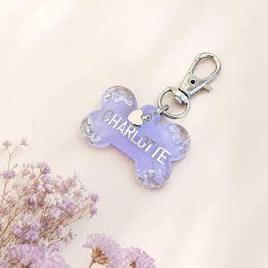 Può includere: Un'etichetta per cani color lavanda a forma di osso con il nome "CHARLOTTE" in bianco. Presenta accenti di foglie d'argento e un fermaglio argentato. È attaccato un piccolo ciondolo a forma di cuore.
