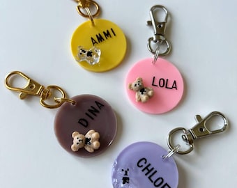 May include: Four colorful round keychains with names: AMMI, LOLA, DINA, and CHLOE. Each keychain has a gold or silver clasp. The keychains are yellow, pink, brown, and purple. Each keychain has a small decorative element.