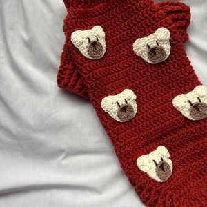 Op de afbeelding: Een handgemaakte, rode gehaakte hondentrui met cr&egrave;mekleurige teddybeergezichten. De trui is gemaakt van een dik, getextureerd garen en heeft een geribbelde kraag. De teddybeergezichten zijn op de trui genaaid.