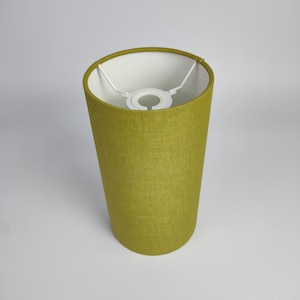 May include: A green cylindrical lampshade made of a textured fabric. The lampshade has a white interior and a white metal ring at the top.