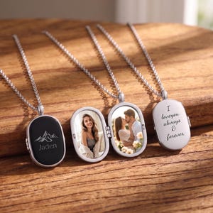 Può includere: Due collane con medaglione ovale e catenine in argento. Un medaglione è chiuso, mostrando un fronte nero con un grafico di montagna e il nome Jaden. L'altro medaglione è aperto, mostrando la foto di una coppia e il testo I love you always & forever.