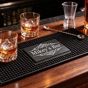Op de afbeelding: Een rechthoekige zwarte rubberen barmat met een getextureerd oppervlak en een centrale witte afbeelding met de tekst "Mikey's Bar" omringd door decoratieve krullen. De mat ligt op een houten oppervlak naast twee kristallen glazen.