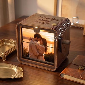 Peut inclure: Une boîte à musique carrée en bois présentant une photo d'un couple s'enlaçant au coucher du soleil. Le dessus est gravé avec le texte I Love You Always et la date 22.06.2026. Une petite manivelle en métal est fixée sur le côté.