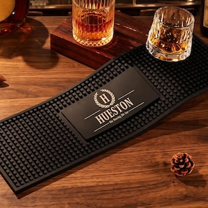 May include: A rectangular black rubber bar mat with a textured surface of small raised nubs. The centre features a smooth rectangular panel printed with a white laurel wreath, the letter H, and the text HUESTON The Roaring 20s bar.