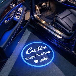 May include: A car door puddle light projector casting a circular blue glow onto the ground. The projection displays the text Custom Image / Text / Logo, a heart icon, and a small car silhouette, illuminating the vehicle interior entrance.