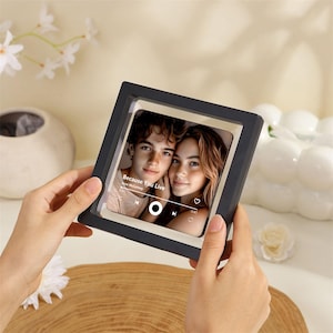 May include: A square black floating frame containing a photograph of a young couple. The image features a music player interface overlay with the text Because You Live by Jesse McCartney, a play button, a heart icon, and a progress bar.