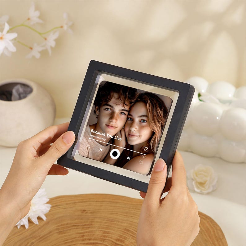 May include: A square black floating frame containing a photograph of a young couple. The image features a music player interface overlay with the text Because You Live by Jesse McCartney, a play button, a heart icon, and a progress bar.