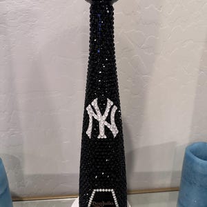 Op de afbeelding: Een zwarte fles bedekt met zwarte strass, met een wit "NY" logo en een honkbalvormige stopper met rode stiksels. De fles heeft de tekst "Don Julio 1942".
