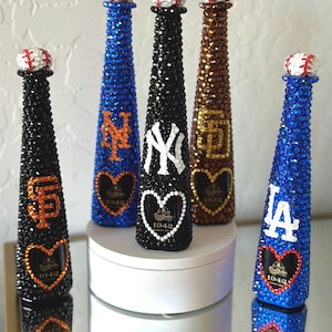 Puede incluir: Cinco botellas de tequila decorativas, cada una adornada con cristales brillantes. Las botellas presentan logotipos de equipos de béisbol, incluidos los Yankees de Nueva York, los Dodgers de Los Ángeles y los Gigantes de San Francisco. Cada botella tiene un tapón en forma de pelota de béisbol.