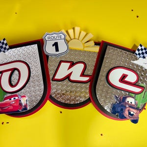 May include: A banner with the word "ONE" in red and white, featuring images of Lightning McQueen and Mater from the movie Cars. The banner has a Route 1 sign and checkered flags, set against a yellow background.