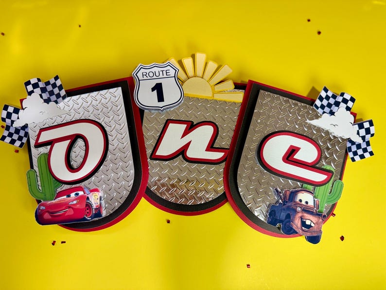 May include: A banner with the word "ONE" in red and white, featuring images of Lightning McQueen and Mater from the movie Cars. The banner has a Route 1 sign and checkered flags, set against a yellow background.