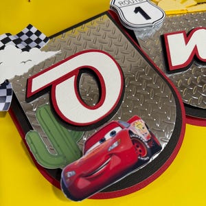 May include: A banner with a red and white "O" and a cartoon image of Lightning McQueen. The banner has a silver diamond plate background, a Route 1 sign, and a cactus. The background is yellow.