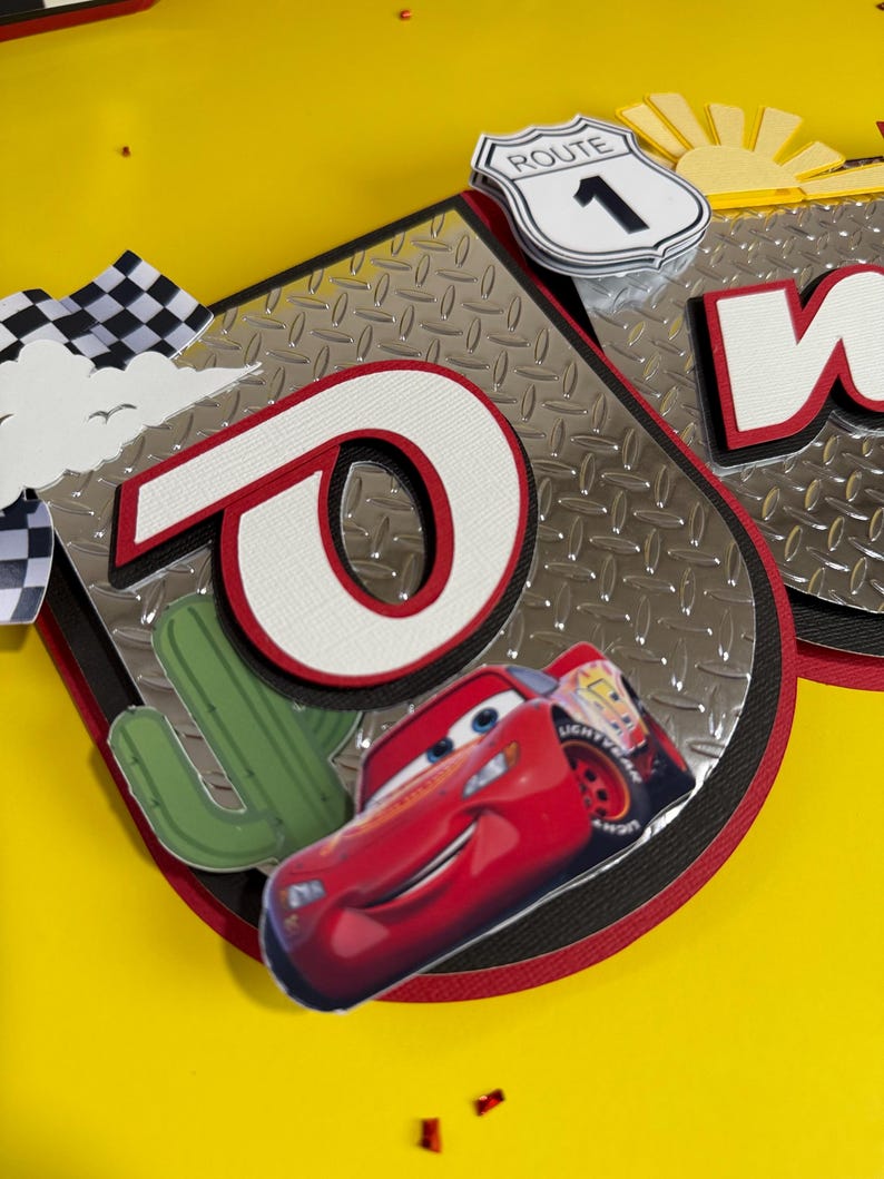 May include: A banner with a red and white "O" and a cartoon image of Lightning McQueen. The banner has a silver diamond plate background, a Route 1 sign, and a cactus. The background is yellow.