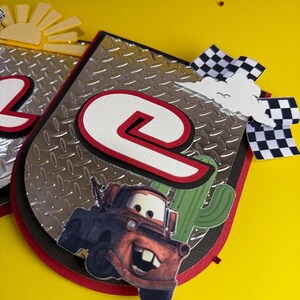 May include: A handmade banner with a race car theme. The banner features a silver diamond plate background, a large number "2" and "9" in red and white, and a cartoon image of a red tow truck. A checkered flag and a cactus are also included.