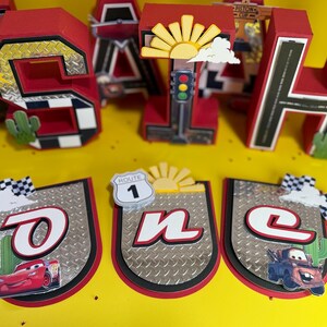 May include: Red and black birthday decorations featuring the word "SAH" and a banner spelling "ONE". The decorations have a race car theme, with images of cars, Route 1 signs, and checkered flags. The letters are approximately 10 inches tall.