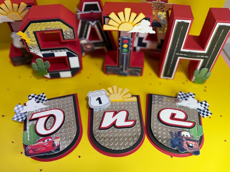 May include: Red and black birthday decorations featuring the word "SAH" and a banner spelling "ONE". The decorations have a race car theme, with images of cars, Route 1 signs, and checkered flags. The letters are approximately 10 inches tall.