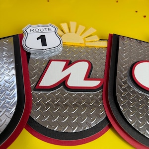 May include: A layered paper craft with a Route 1 sign, a yellow sun, and the letter "n" in white with a red outline. The background is a yellow surface. The design incorporates silver diamond plate patterns and black and red borders.