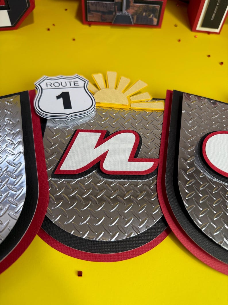 May include: A layered paper craft with a Route 1 sign, a yellow sun, and the letter "n" in white with a red outline. The background is a yellow surface. The design incorporates silver diamond plate patterns and black and red borders.